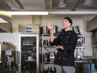 The image shows a chef standing confidently in a professional commercial kitchen. He is wearing a black chef’s uniform and a matching hat, with a thoughtful or expressive hand gesture, as if evaluating a dish or emphasizing a culinary idea.