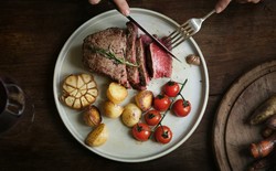 A plate with roasted meat, potatoes, fresh tomatoes, and garlic, with a glass of wine next to it