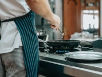 The image shows a chef cooking on a stovetop in what appears to be a restaurant or front-line kitchen. The chef is wearing a striped apron and checkered pants, indicating a professional culinary environment.