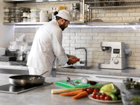 A chef wearing a white toque and uniform is working in a brightly lit, modern kitchen. He is standing at a stainless steel sink, washing some orange vegetables