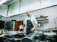 The image shows a smiling chef working in a professional kitchen. He is wearing a white chef's jacket and a dark striped apron. The chef is cooking at a gas stove, with several pots and pans in use.