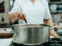 The image shows a chef in a professional kitchen stirring a large stainless steel pot on a stovetop.