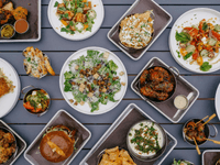 The image displays a vibrant and appetizing spread of various dishes arranged on a dark gray table. The food appears to be from a modern American or fusion-style restaurant.
