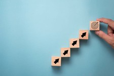 This image shows a hand placing a block with a target icon at the top of a staircase-like arrangement of wooden blocks, each marked with an arrow pointing upward. It symbolizes progression or steps toward a goal, often used in business or strategy contexts to represent growth, development, or goal-setting processes.
