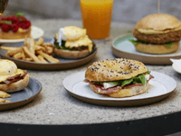The image showcases a variety of well-presented gourmet dishes — including a deli-style bagel, eggs benedict, artisan burger, fries, and fresh juice — designed to convey quality, variety, and professionalism. The clean, polished setup reflects the brand’s ability to cater to corporate events, hotels, cafes, and businesses looking for reliable food service partners.