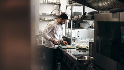 Chef in a modern dark kitchen prepares food for delivery, cutting ingredients on a vibrant green cutting board. The space is filled with industrial-grade appliances, utensils, and plates.