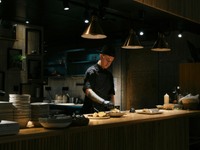 This is a photograph of a chef working at the pass or service counter of a kitchen with a modern, dark design aesthetic.