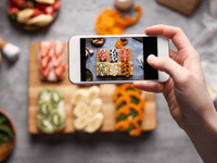 A person is taking a photo of colorful, neatly arranged fruit and vegetable snacks using a smartphone. The vibrant ingredients are creatively displayed on a wooden board, making the setup highly Instagrammable.
