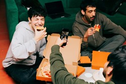 Friends enjoying a casual meal from a food delivery service, gathered around a green couch with pizza boxes and slices of pizza in hand. One person captures the moment by taking a photo with their smartphone.