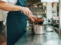 The image shows a chef in a professional kitchen serving food from a stainless steel pot into a small dish using a ladle. The chef is wearing a striped apron, indicating a food service or culinary professional.
