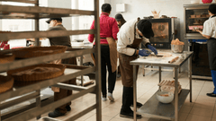 The image shows a busy bakery or pastry kitchen with several workers preparing baked goods.