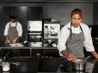 This photograph features a female chef actively working in a modern, professional kitchen, with a colleague preparing food in the background.