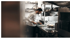 A professional chef wearing a white jacket stands in a modern commercial kitchen, carefully chopping ingredients on a cutting board.