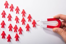 This image depicts a hand holding a magnet attracting small red figurines shaped like people. It symbolizes customer attraction or lead generation, often used in marketing or business contexts to represent strategies for drawing in clients or users.