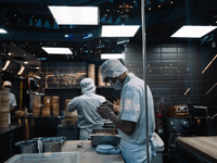 This photo, taken through a glass window, shows three chefs working in a busy, modern kitchen. The glass creates reflections of the ceiling lights and surrounding area.