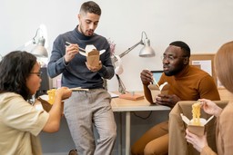 A group of young people sharing a meal with Asian food in typical packaging, in a modern office setting.