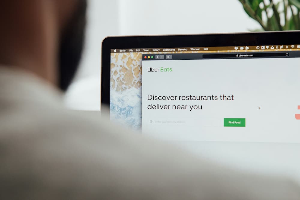 A person views the Uber Eats website on a laptop screen. The website shows the Uber Eats logo along with a search bar and the message 'Discover restaurants that deliver near you.'