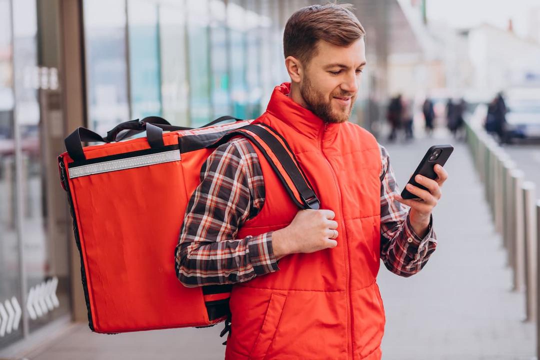 Photograph of a delivery rider with a food delivery bag, looking at a smartphone. Photo: Freepik.