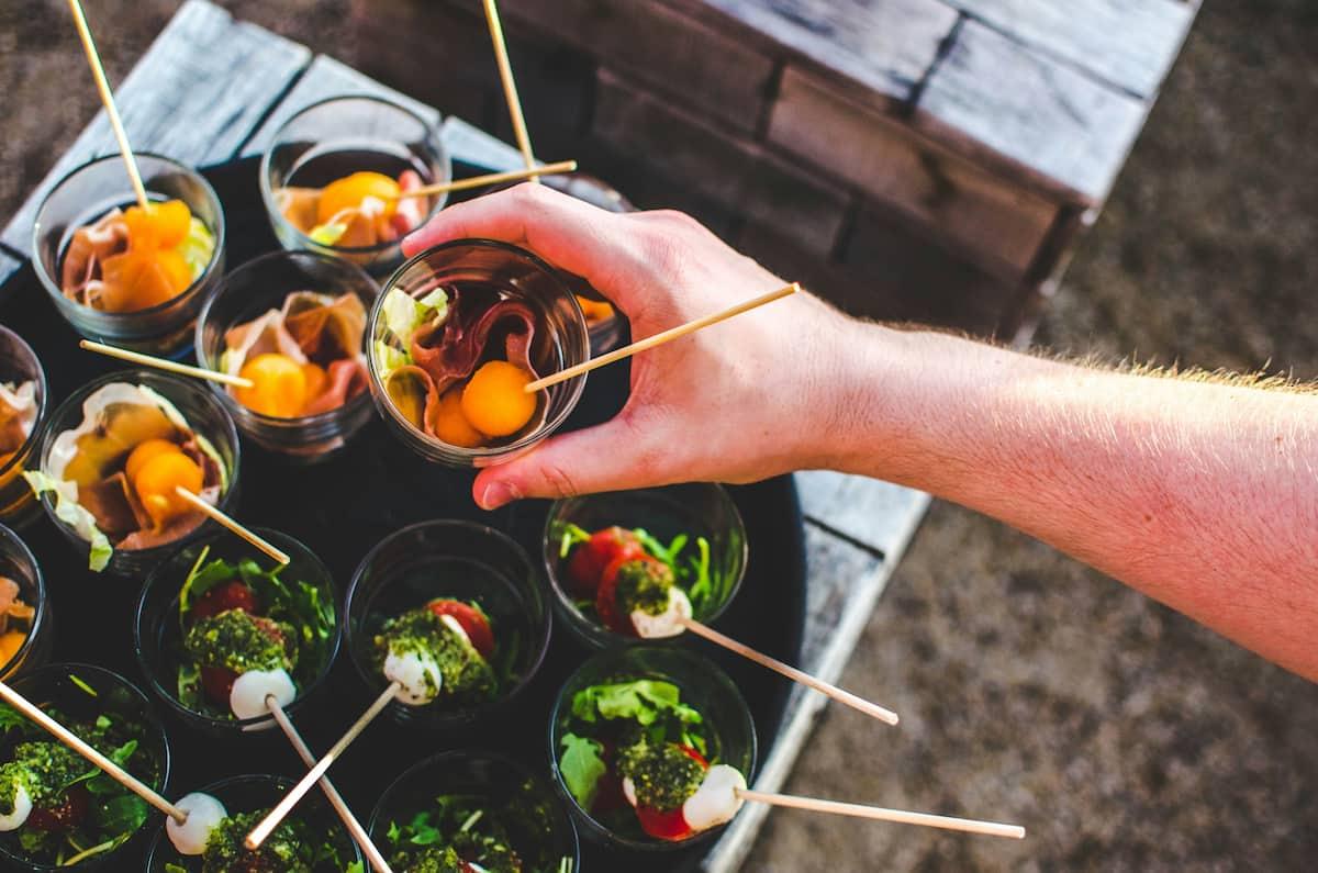 Hand holding a glass cup with melon balls and prosciutto skewers, with similar appetizer cups in the background.