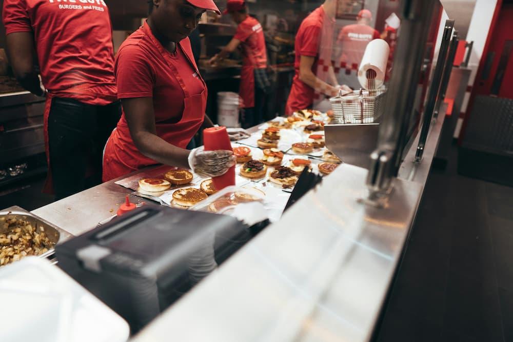 Fast food restaurant kitchen with employees preparing burgers. Workers in red uniforms and hats are assembling burgers with various toppings on a stainless steel counter.