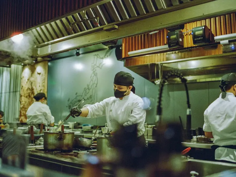 The image shows a professional kitchen in full activity. In the center, a chef wearing a white uniform, black hat, and black mask is preparing a dish in a large pot on the stove.