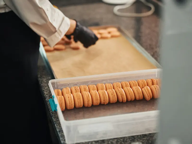 This close-up photo shows someone's hands, wearing black gloves, working on a large batch of macaron cookies in a kitchen.