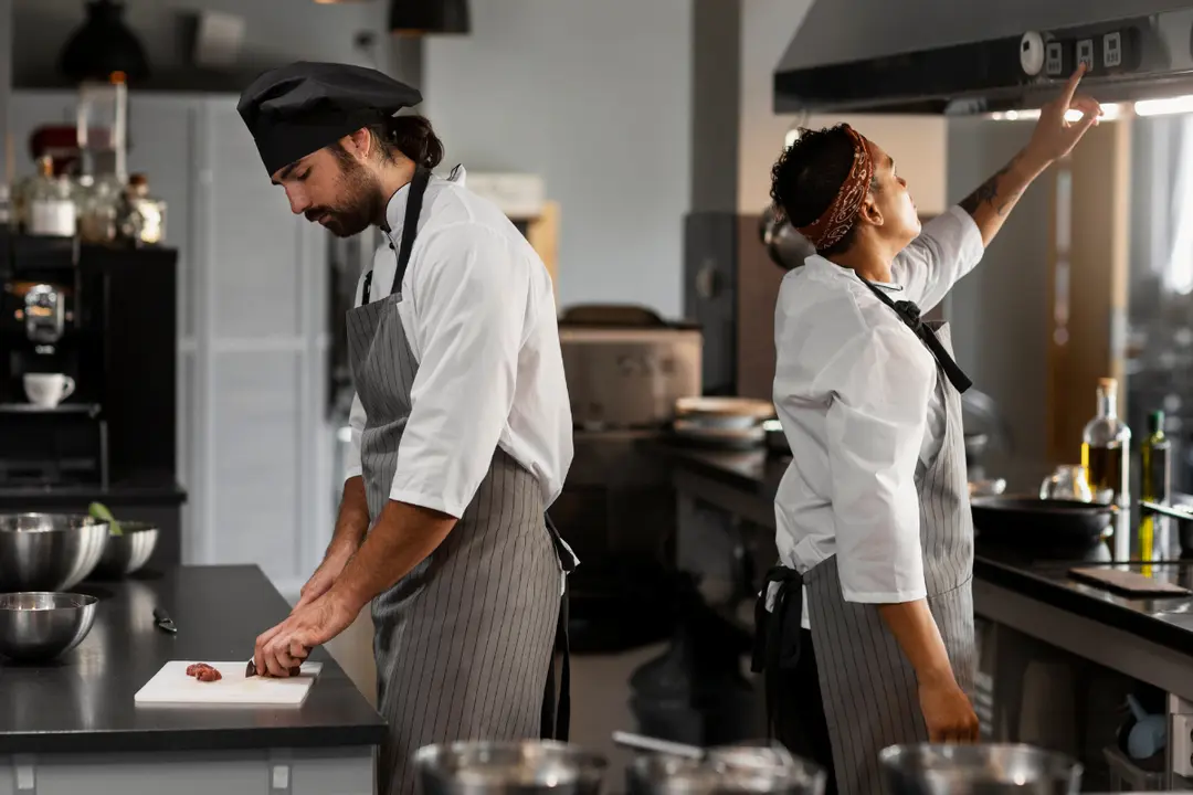 This photograph shows two professional chefs working side-by-side in a commercial kitchen, engaged in different tasks.