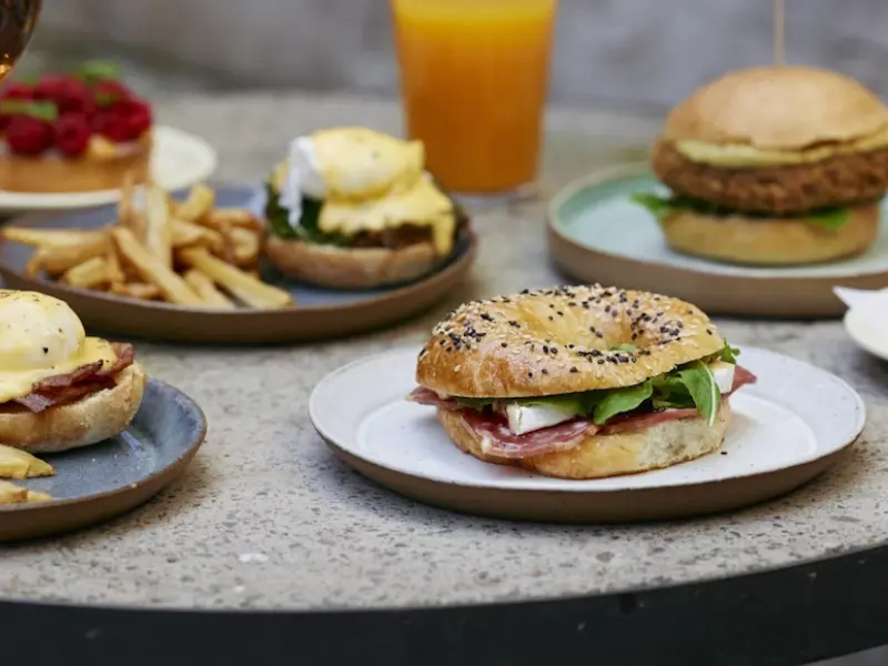 The image showcases a variety of well-presented gourmet dishes — including a deli-style bagel, eggs benedict, artisan burger, fries, and fresh juice — designed to convey quality, variety, and professionalism. The clean, polished setup reflects the brand’s ability to cater to corporate events, hotels, cafes, and businesses looking for reliable food service partners.