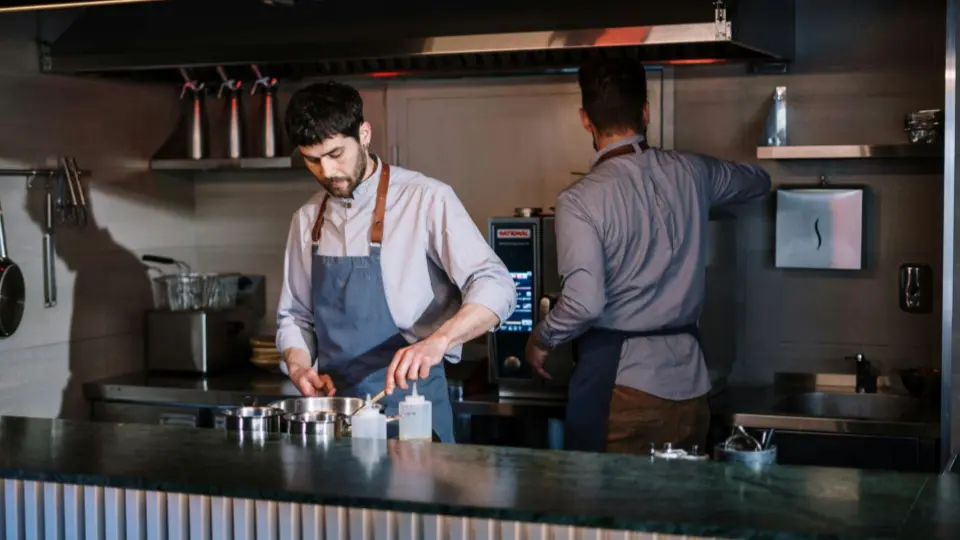 The image shows two chefs working in a professional, industrial-style kitchen.