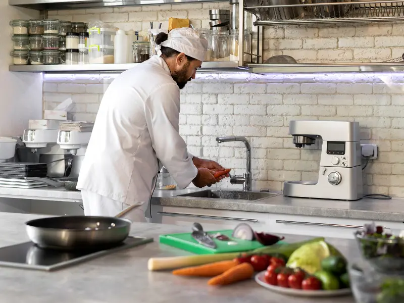 A chef wearing a white toque and uniform is working in a brightly lit, modern kitchen. He is standing at a stainless steel sink, washing some orange vegetables