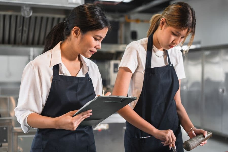The image shows two women working in a kitchen, wearing white shirts under black aprons. One is holding a clipboard, seemingly taking notes or inspecting, while the other is focused on preparing food with a utensil. The setting appears to be a professional kitchen, possibly a restaurant or food service establishment, with stainless steel equipment visible in the background.