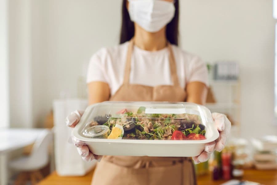 This image shows a person holding a packaged food container, wearing a face mask and gloves, suggesting a food preparation or delivery environment, possibly related to meal delivery services or ghost kitchens, emphasizing safety and hygiene in food handling.