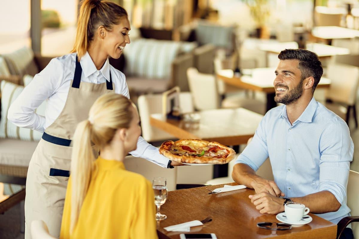 A waitress is serving a freshly baked pizza to a smiling couple seated at a restaurant table, fostering a warm and friendly atmosphere.